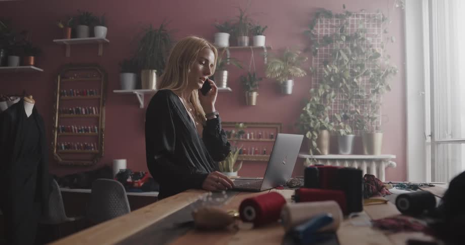 A woman is standing at a table typing on a laptop computer and she answers her cell phone