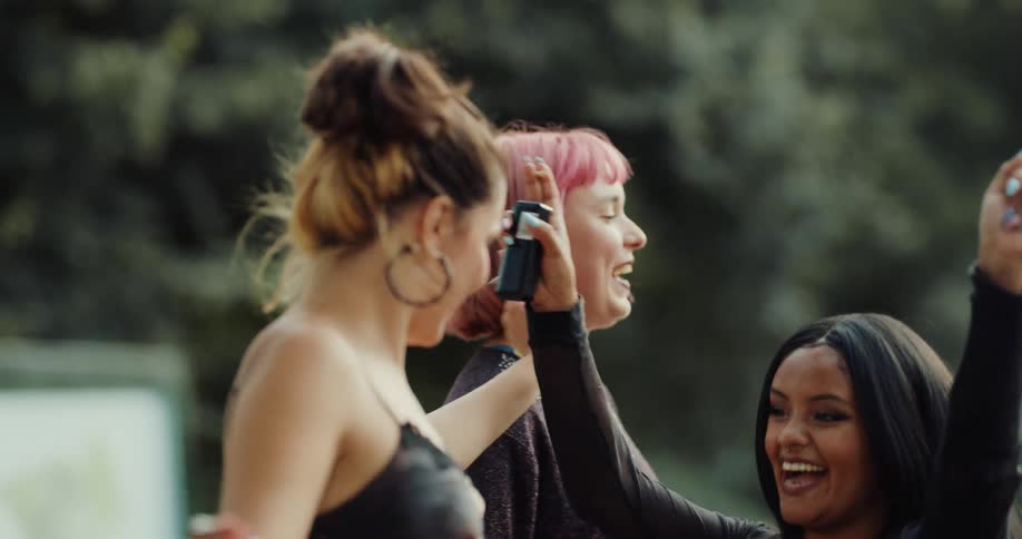 Group of young female friends having fun together dancing and playing a musical instrument in a public park on a sunny day