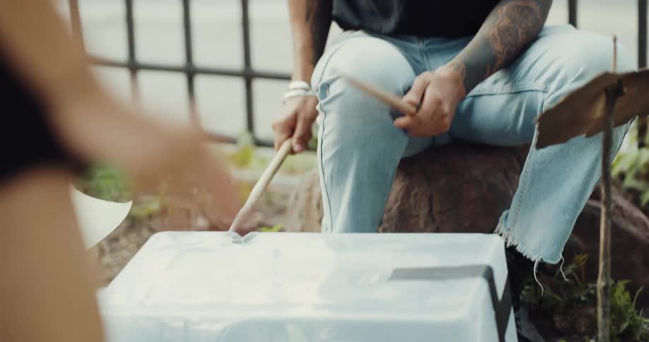 A male is sitting outdoors and playing the drums with his friend