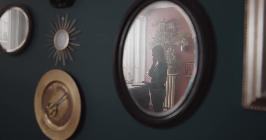 Mirror reflection of a woman standing in front of a counter using her laptop and talking on the phone at the same time