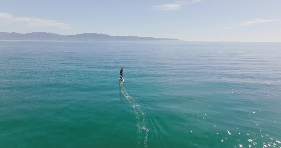 A woman rides an electric hydrofoil surfboard across sparkling clear blue water and there are mountains in the distance