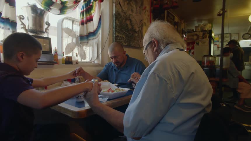 A family of three generations of men holding hands and praying before eating at a restaurant