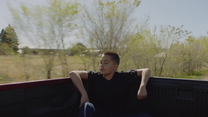 A boy is riding in the back of a pickup truck as it travels on a rural road
