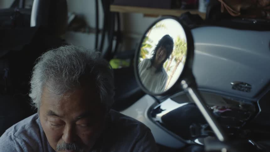 A older man is repairing a motorcycle in the garage with the reflection of a younger man in the mirror
