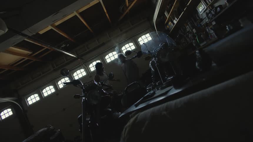 Two men share a smoke while standing in dimly lit garage near motorcycles
