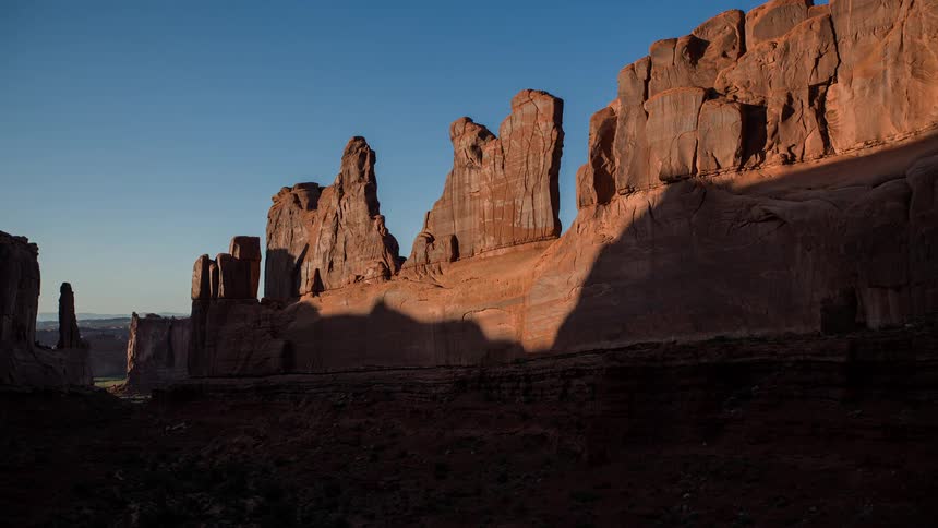 A shadow moves across red rock formations in the desert under a clear blue sky
