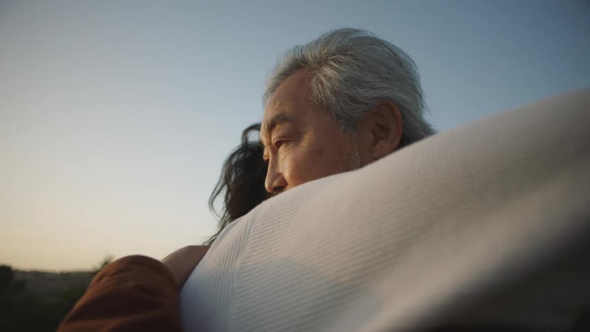 A man closes his eyes while hugging his adult son outside at sunset