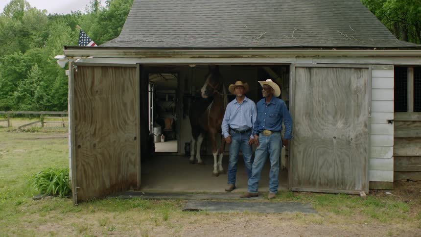 Two older cowboys stand at the front of a barn near horse on farm in countryside