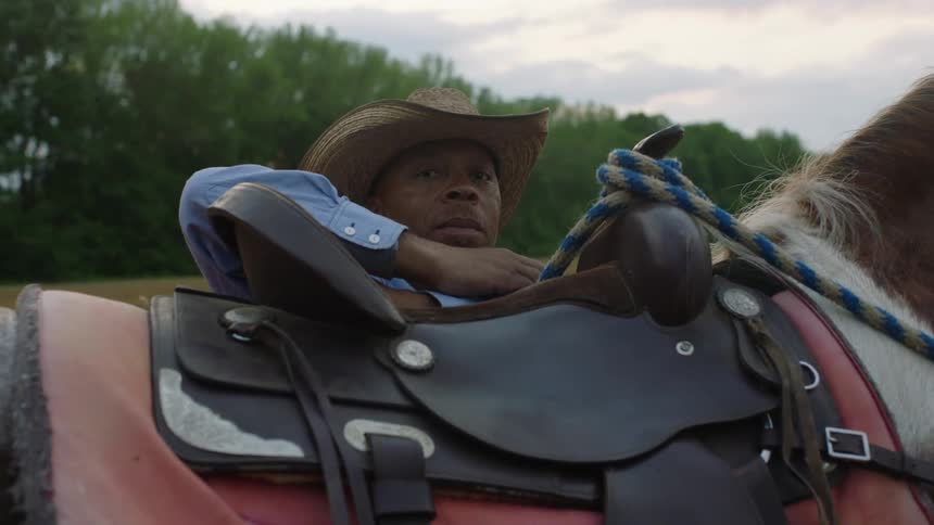 A man standing behind a saddled horse with his arm leaning on the saddle