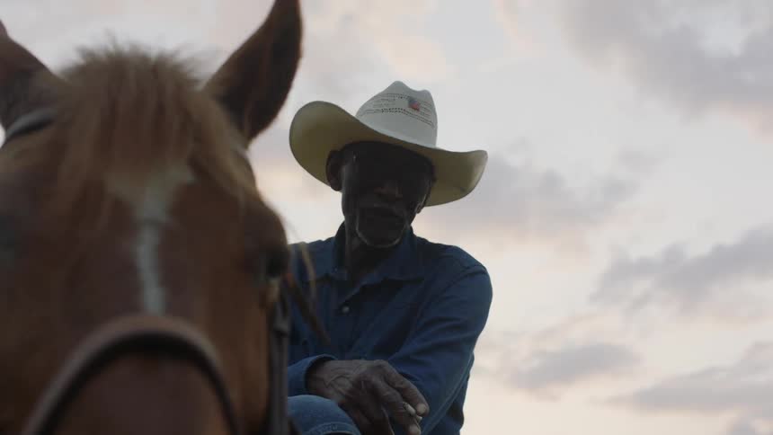 Black man wearing a cowboy hat holding a cigarette while sitting on brown horse