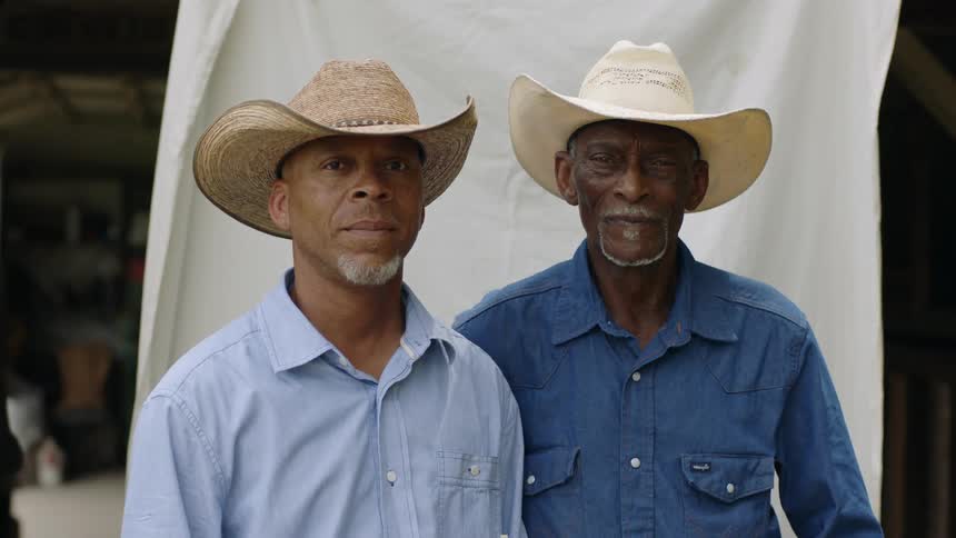 Two men wearing cowboy hats standing in front of a stable looking at the camera