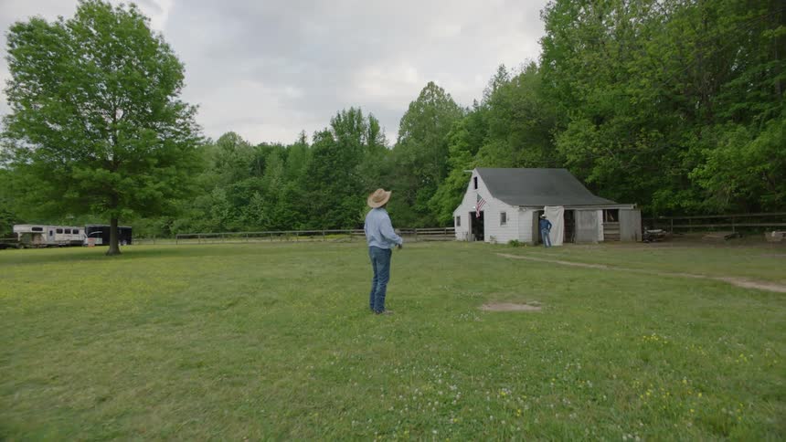 A man wearing a cowboy hat is standing in the yard facing the barn where another man wearing a cowboy hat is standing