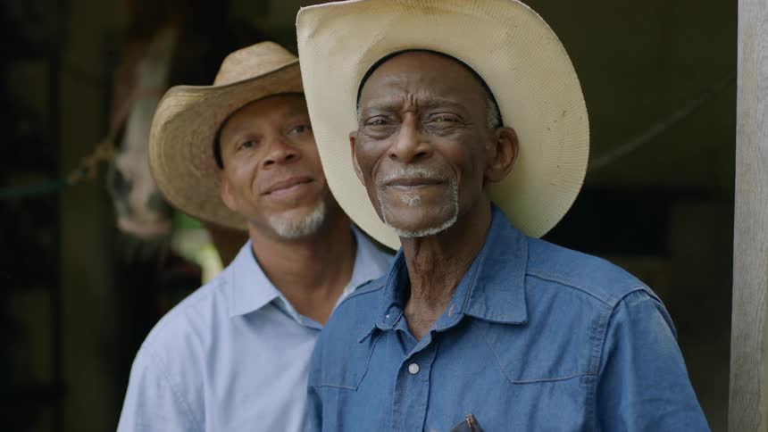 Two men wearing cowboy hats standing in front of a stable looking at the camera