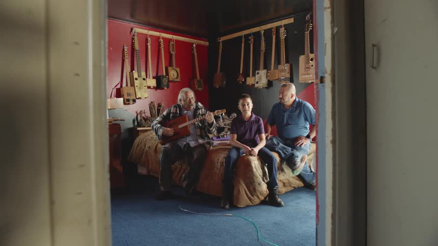 Family sits on the bed on a bedroom at home while grandpa plays guitar
