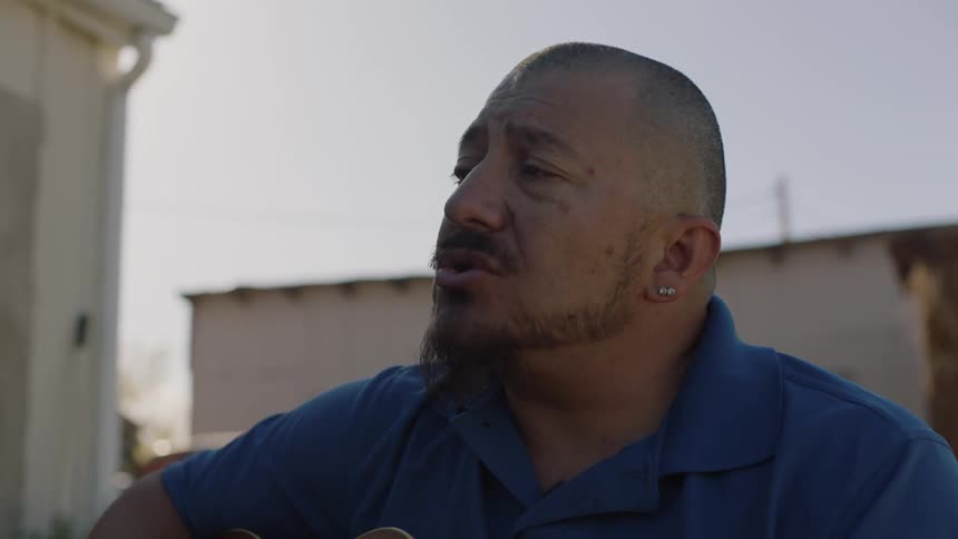 Man sits outdoors near door to a building singing and strumming a guitar