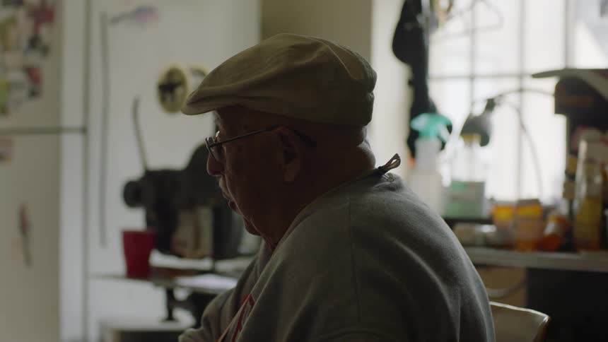 A elderly man sits at his kitchen table by the window