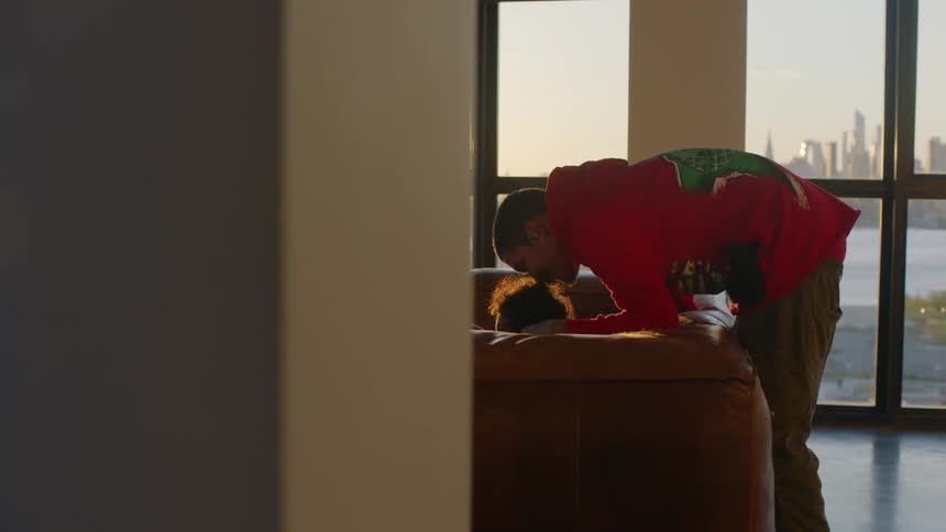 Father bends over couch and child as large windows show water and skyline