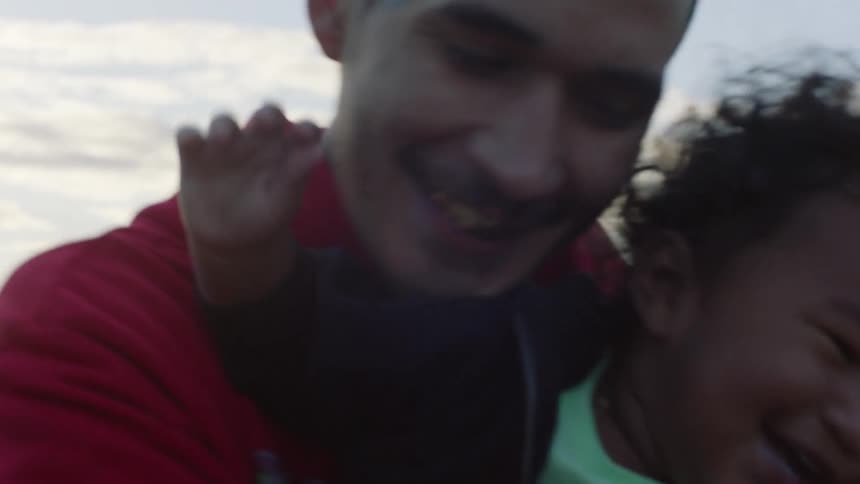 Father smiles while hugging laughing child on lake shore opposite city skyline