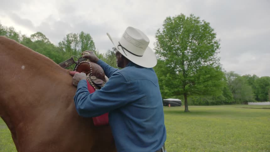 Black man wearing a cowboy hat putting a saddle on the back of a horse in the middle of a grass field on a farm