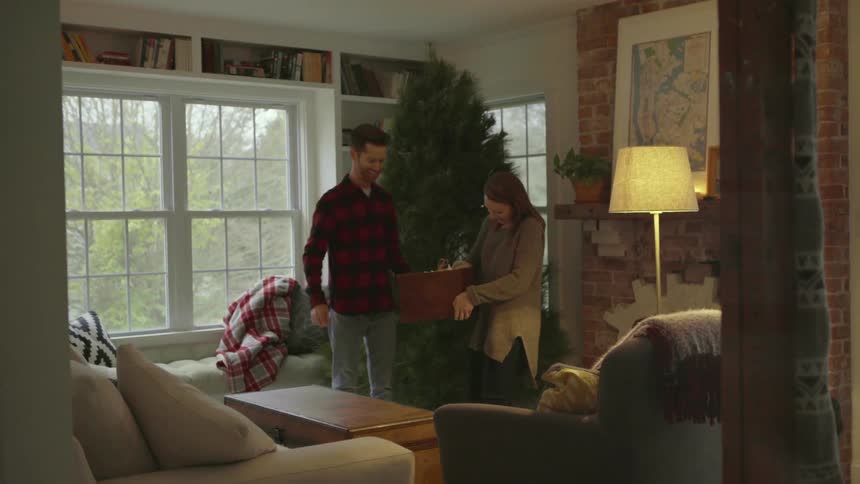 A woman is carrying a box of ornaments and sits them on a table next to a empty christmas tree