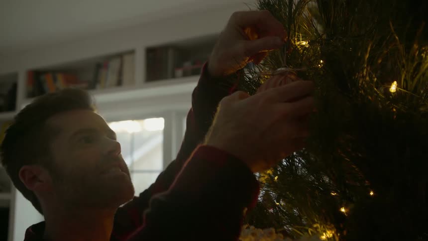 A man hangs an ornament on a christmas tree then steps back