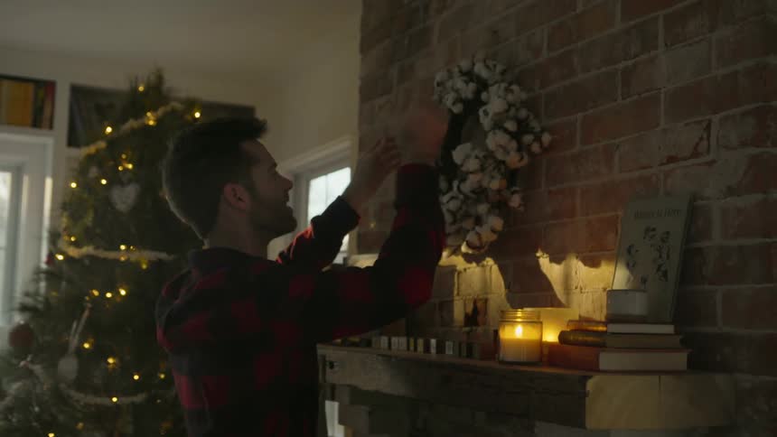 A man is hanging a Christmas wreath above a fireplace