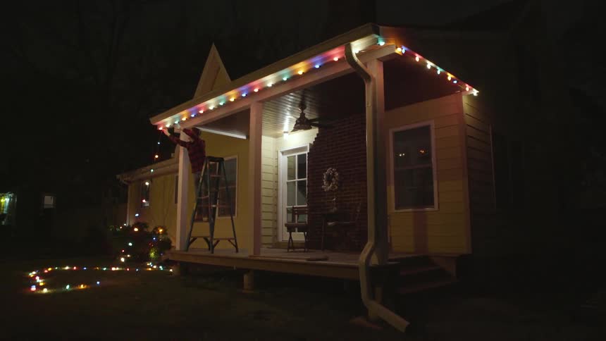 Man on a ladder stringing christmas lights on his house at night