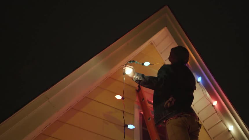 A man is hanging christmas lights from the roof