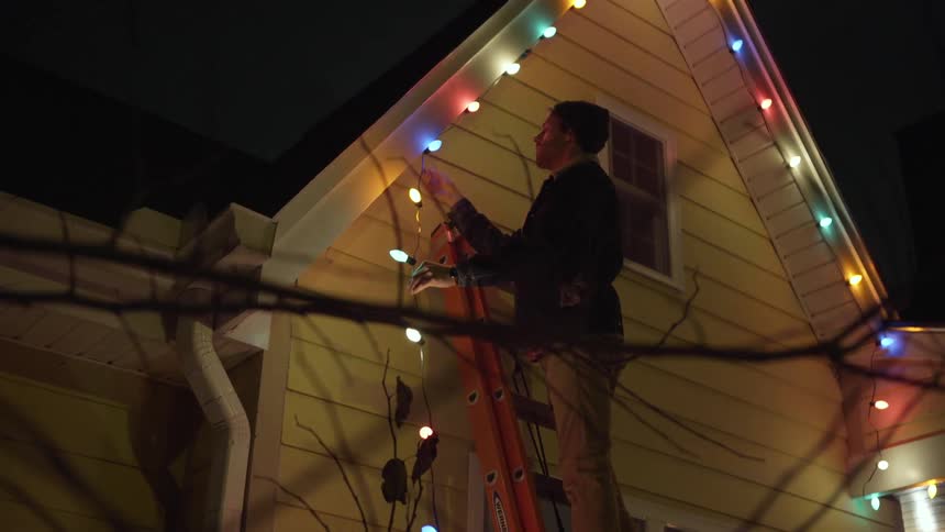 A man climbs up a ladder and hangs christmas lights on his house