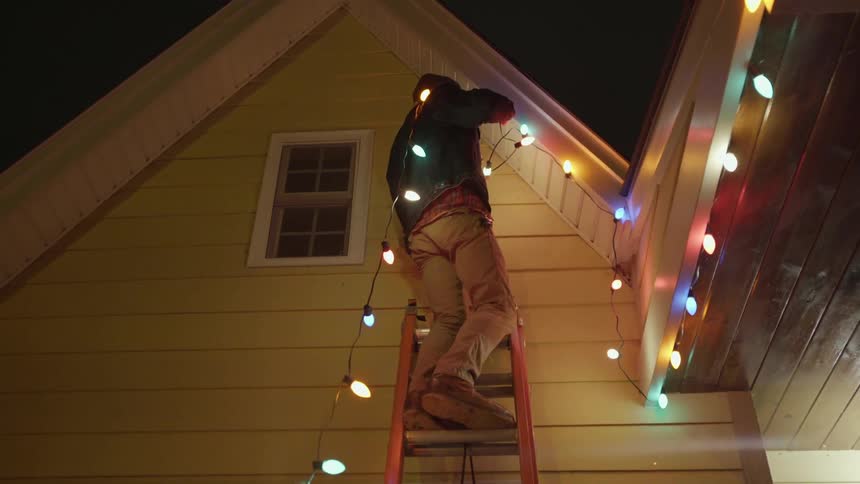 A man is climbing a ladder with christmas lights on his shoulder
