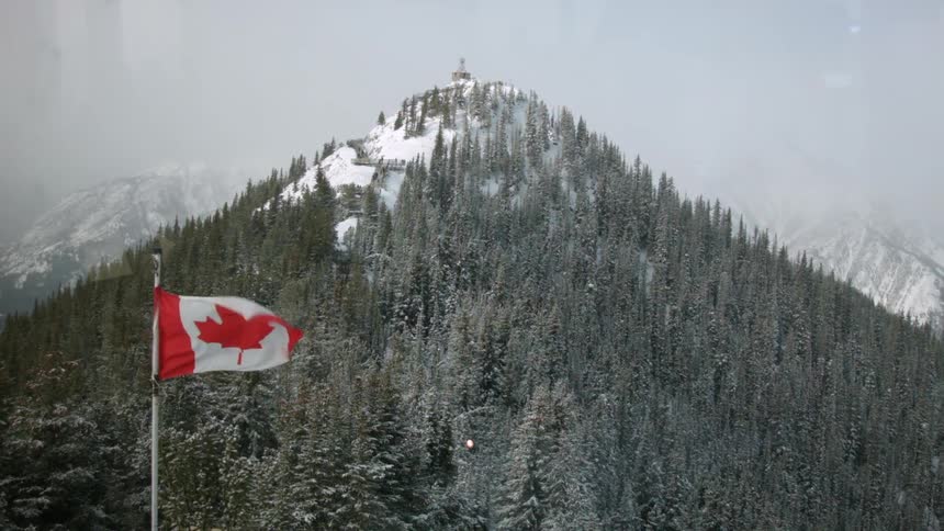 The canadian flag waving in the breeze in front of a forested and snow covered mountain