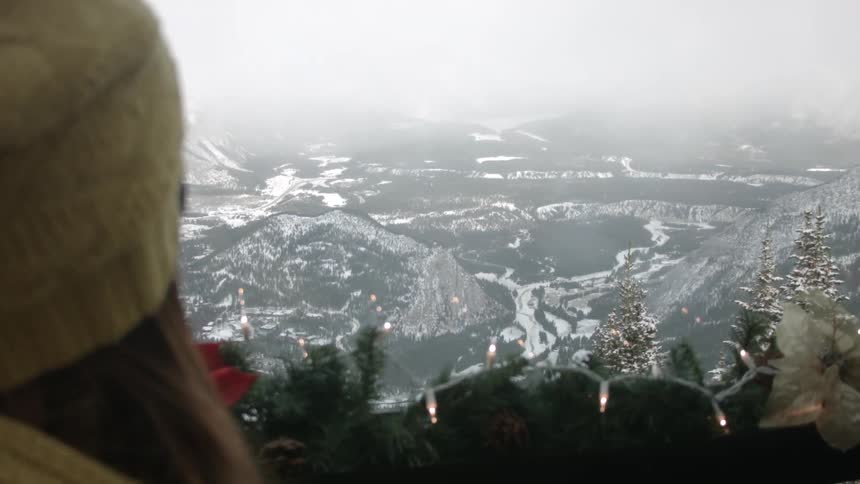 A woman sitting on a deck views the landscape far below in the valley