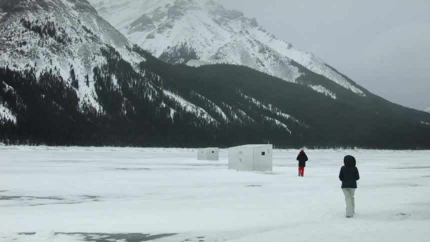Two people walk across a frozen lake towards fishing huts with snow covered mountains in the background