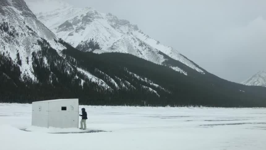 A woman approaches a small utility building located on a snowy plain at the base of snow-capped mountains