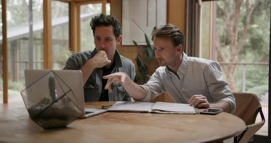 Two men are sitting at a table, one is using a laptop and the other is looking at paperwork