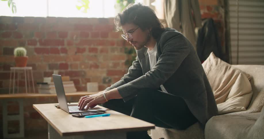 A man leans forward as he sits on a couch and types on a laptop on the coffee table
