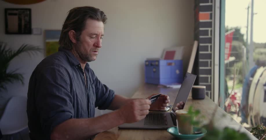 A man sitting at a counter is inputting credit card information into a laptop