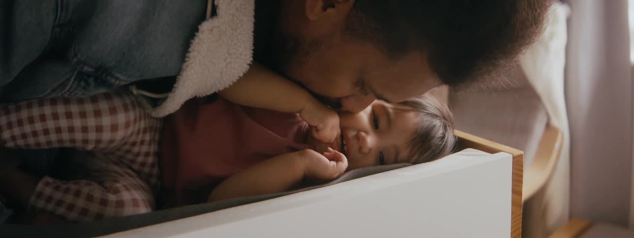 Father is kissing talking and playing with his young daughter who is lying down in the cradle