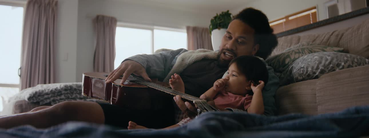 A father plays guitar with his young daughter sitting on the couch next to him