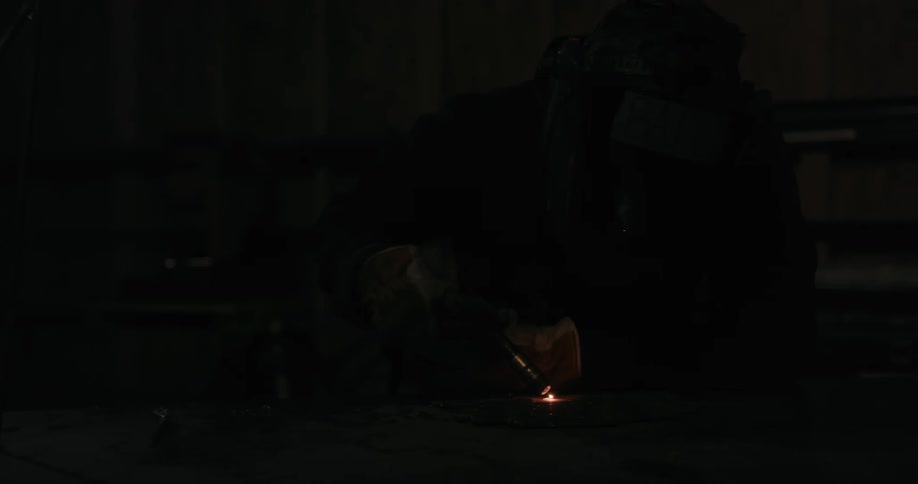 A welder leaning over working on metal causing smoke and sparks at night