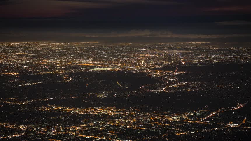 Plane and traffic lights flow across brightly lit cityscape after sun sets