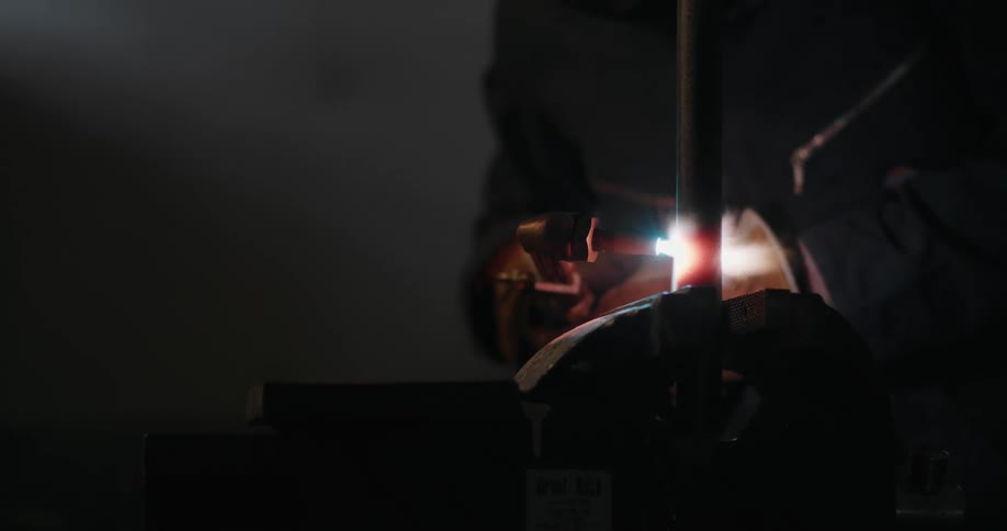 Welder uses torch on metal pole clamped in vise in dim room
