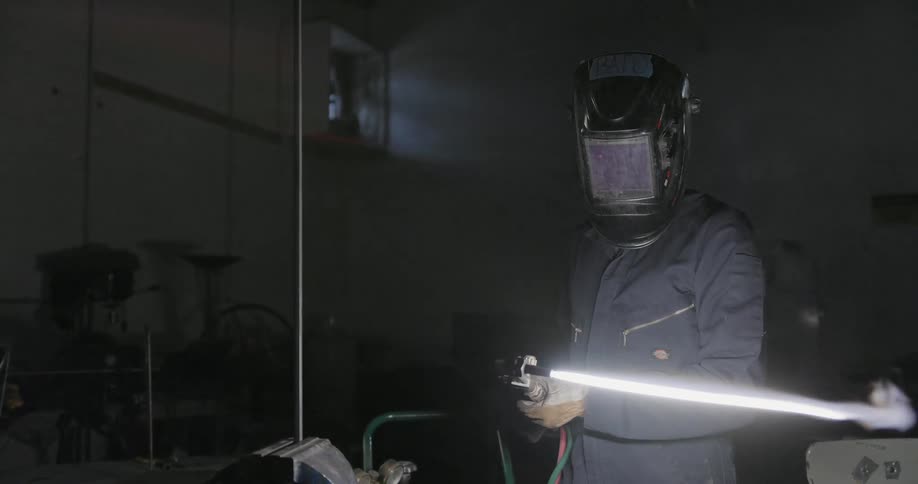 Man puts down welders mask as he stands in dim room holding burning blowtorch