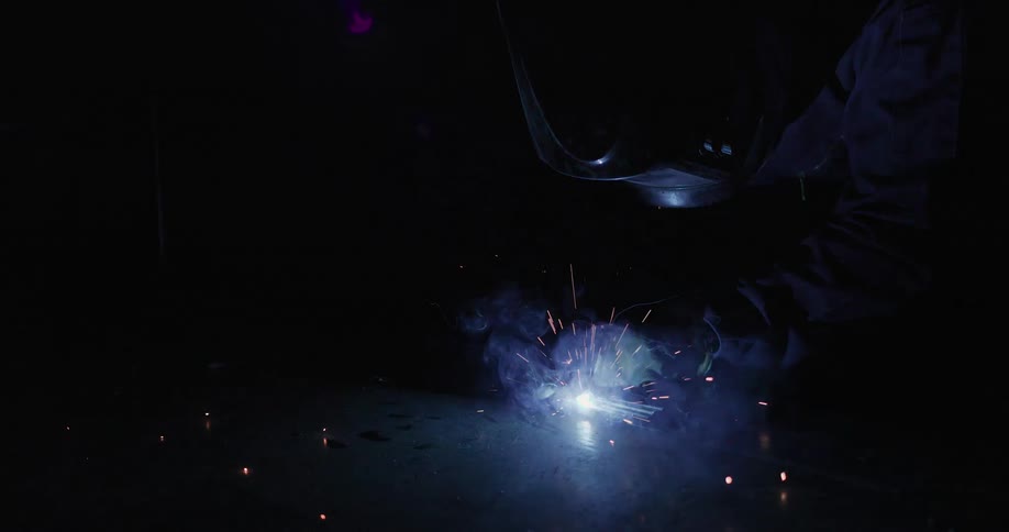 Sparks fly and shine brightly as smoke rises in the darkness as someone wearing a helmet is welding
