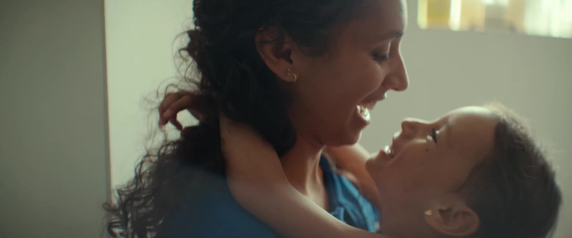 A little girl with her hands around moms neck smiling up at her while mom kisses her