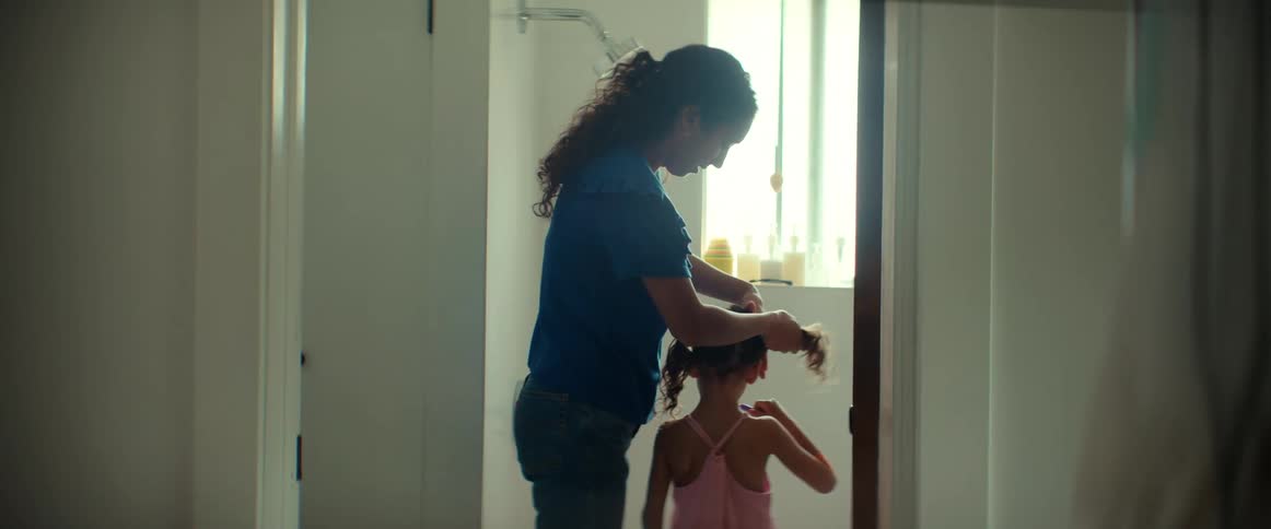 Mom puts her daughters hair into pigtails while they both stand in the bathroom near a window