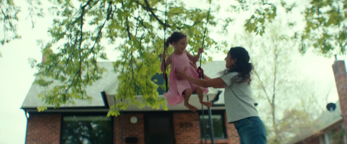 Mother pushes daughter in swing attached to tree branch in front of house