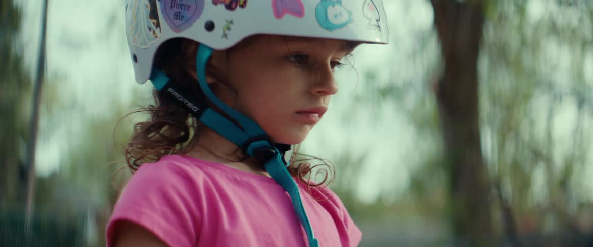 A little girl wearing a helmet is standing outside near trees and frowning