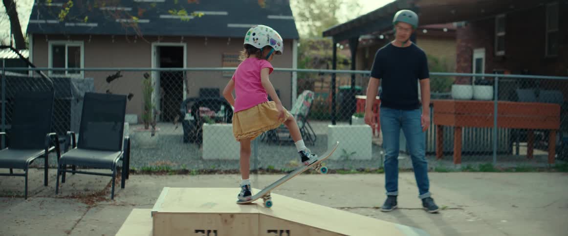 A little girl wearing a helmet rides down a ramp on a skateboard in her neighborhood as her father watches