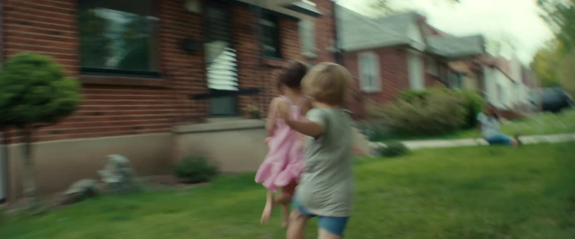 A little boy chasing a little girl through the grass of a their front yard as mom watches